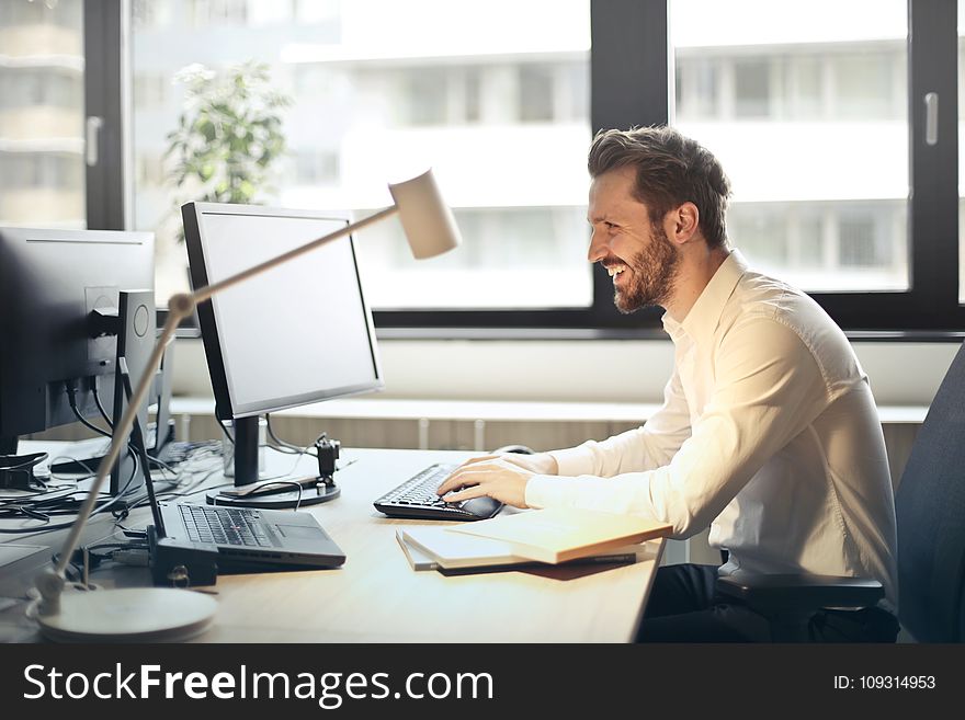 Man in White Dress Shirt Sitting on Black Rolling Chair While Facing Black Computer Set and Smiling