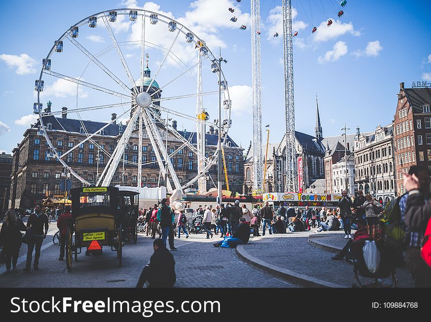 Ferris Wheel Beside Brown Buildings and People Under Blue Cloudy Sky at Daytime