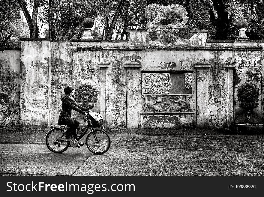 Woman Wearing Jacket and Pants Riding on Bicycle Near Concrete Wall Greyscale Photo