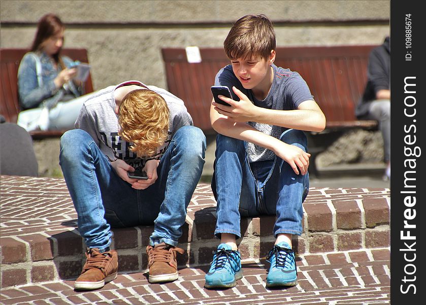 2 Boy Sitting On Brown Floor While Using Their Smartphone Near Woman Siiting On Bench Using Smartphone During Daytime