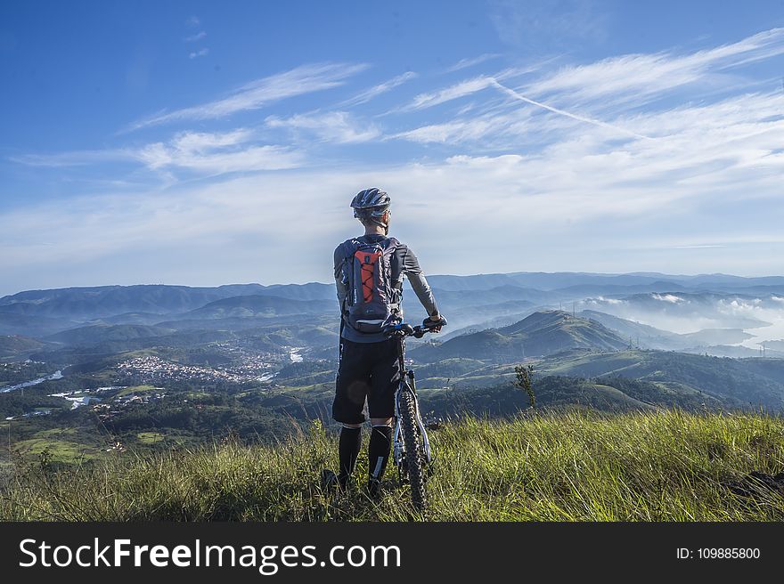 Biker Holding Mountain Bike On Top Of Mountain With Green Grass