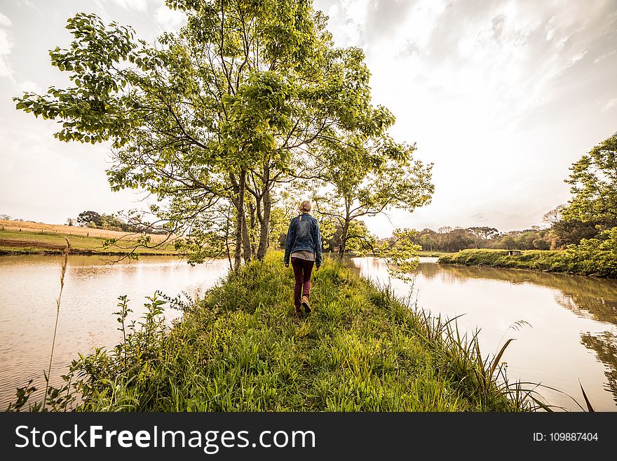 Person In Blue Denim Jacket And Brown Pants Standing On Green Grass In Front Green Leaved Trees Between River Under Sunny Sky