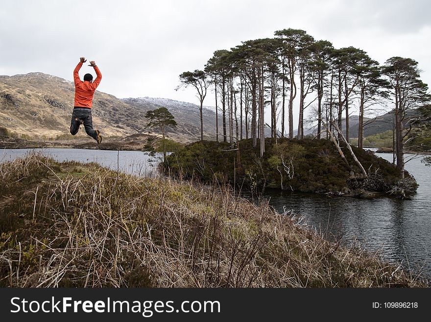 Man in Orange Long-sleeved Shirt Jumping on Lake Near Tall Trees at Daytime