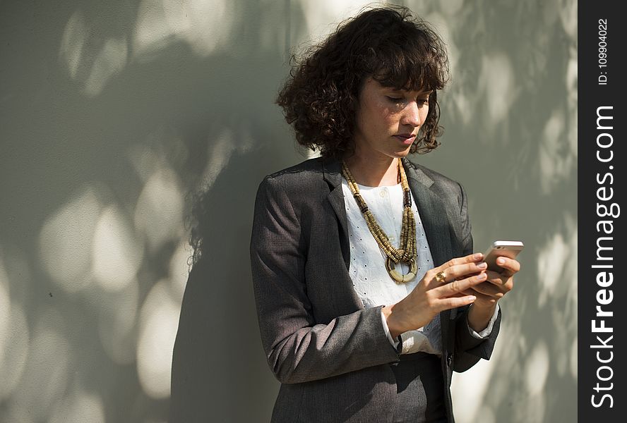 Woman In Black Blazer Holding A Smartphone While Standing Near Wall