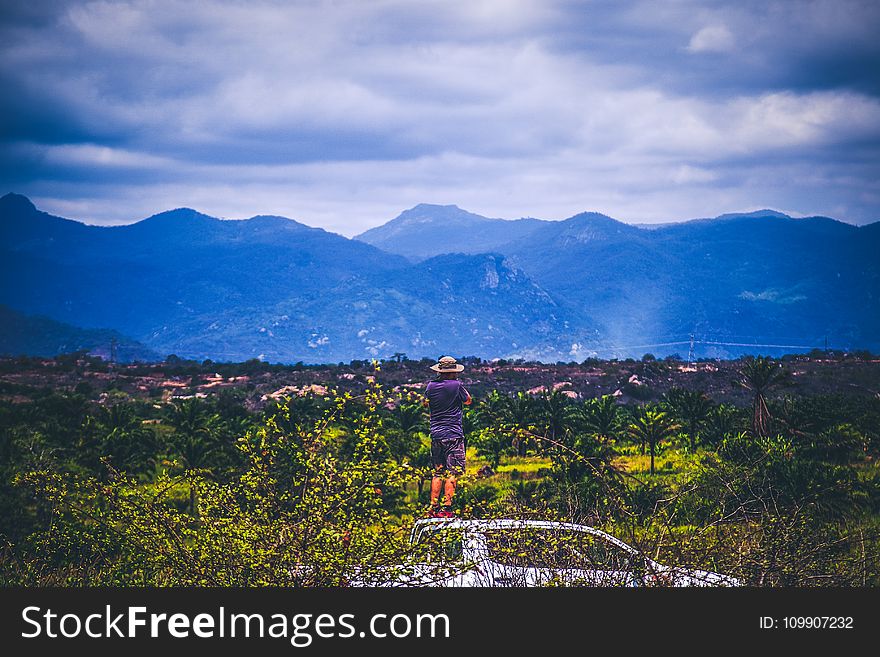 Person Wearing Sun Hat Looking At Mountains Under Cloudy Sky