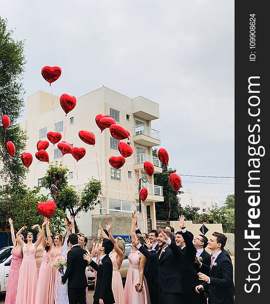 Women Wearing Pink Dresses and Men Wearing Black Suit Jacket and Pants Raising Hands With Red Heart Balloons