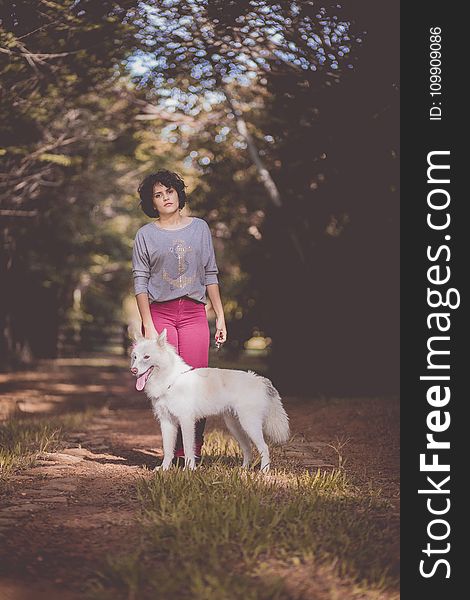 Woman in Purple Long Sleeve Shirt and Pink Jeans Standing Next to White German Spitz With Background of Green Trees during Day