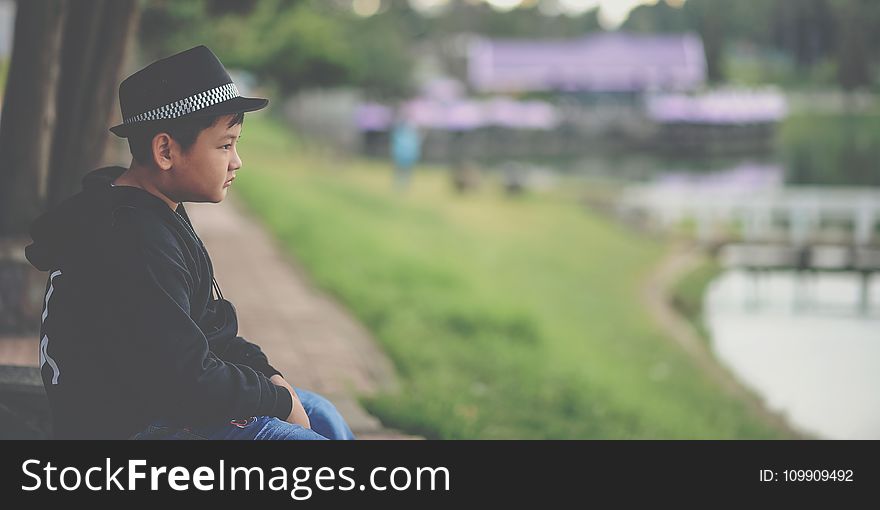 Boy in Black Jacket and Black Fedora Hat Sitting Near Body of Water