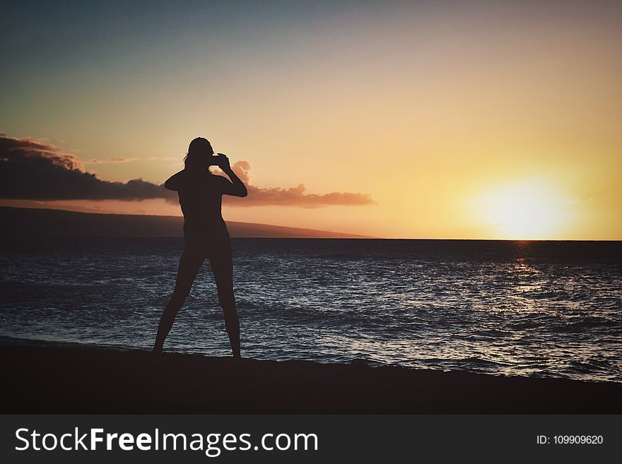 Silhouette of Woman Holding Camera Near Seashore during Golden Hour