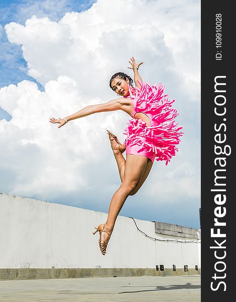 Woman In Pink Dress Doing Jump Shot While Extending Arms Under White Clouds