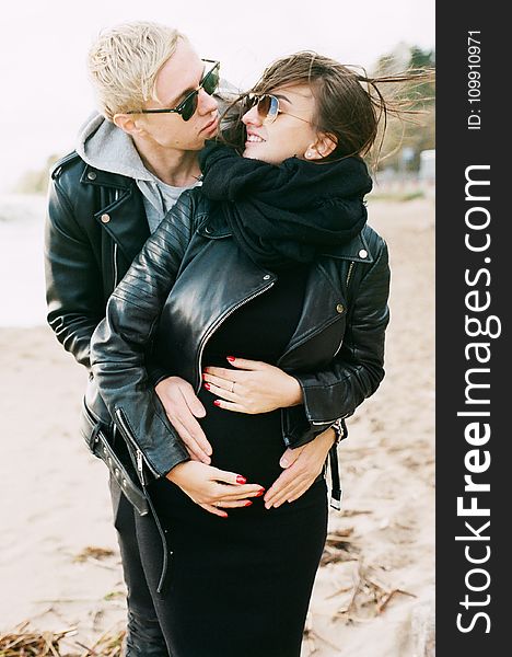 Man in Black Leather Zip Jacket Hugging Woman in Black Leather Zip Jacket and Black Pants Standing on Sand at Daytime