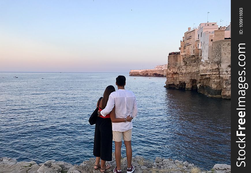 Man in White Dress Shirt Standing Beside the Woman in Black and Red Dress While Watching the Blue Calm Water Near Brown Concrete Buildings Under White and Blue Sky at Daytime