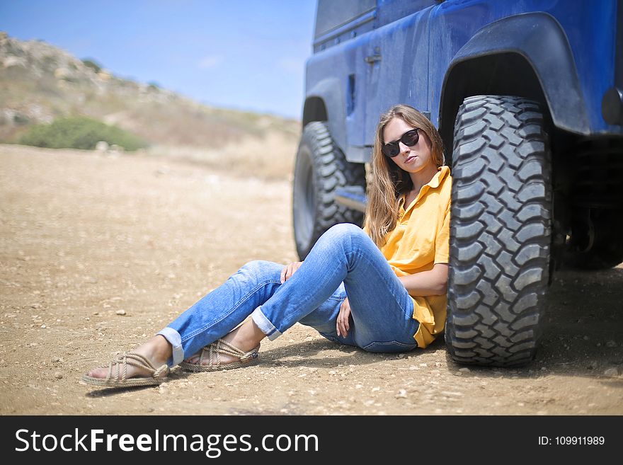 Woman in Yellow Polo Shirt Sitting on Ground Leaning on Blue Vehicle at Daytime