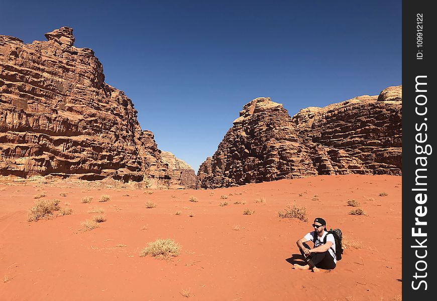 Man Wearing White Shirt and Black Pants Sitting on Soil Behind Rock Formation Mountain