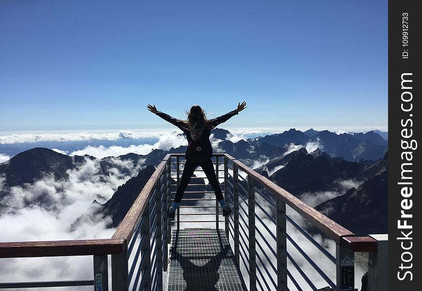 Person Standing on Hand Rails With Arms Wide Open Facing the Mountains and Clouds