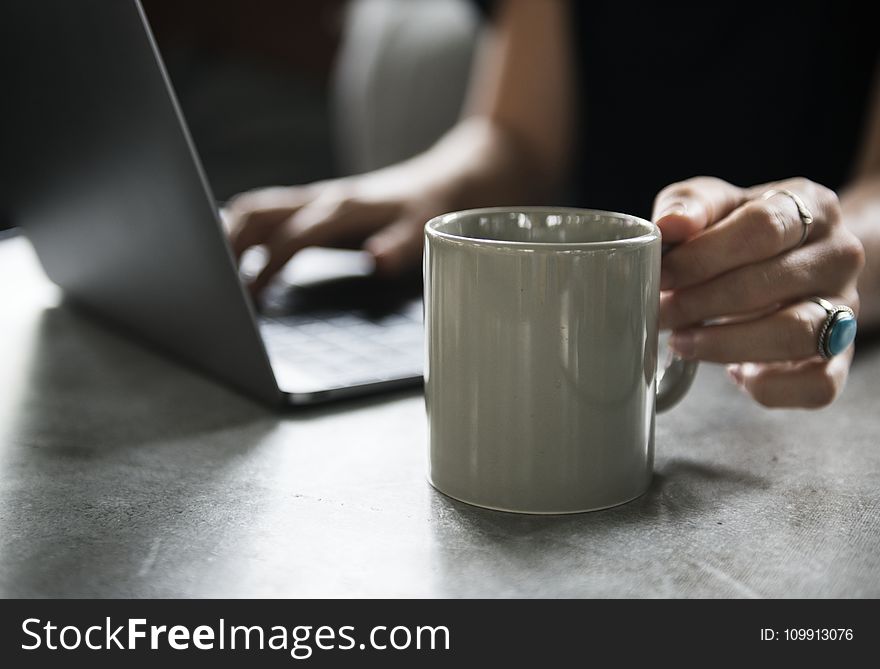 Person in Black Top Holding White Ceramic Mug and Using Laptop Computer