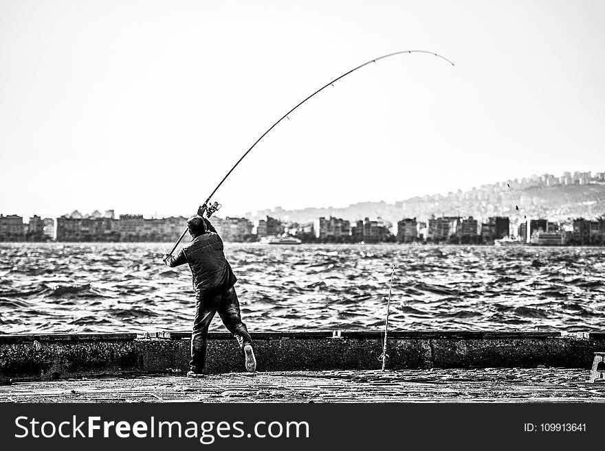 Grayscale Photography of Man Holding a Fishing Rod Near Body of Water