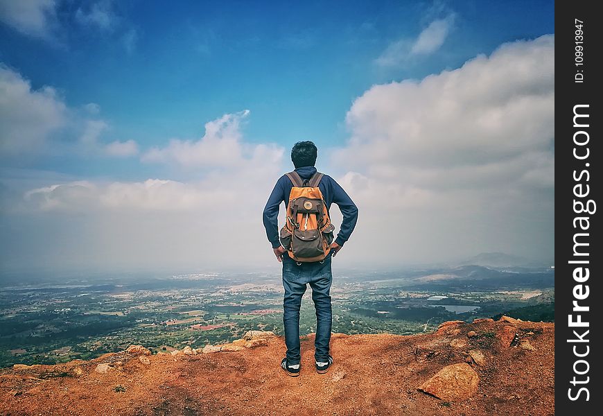 Man in Blue Dress Shirt and Blue Jeans and Orange Backpack Standing on Mountain Cliff Looking at Town Under Blue Sky and White Clouds