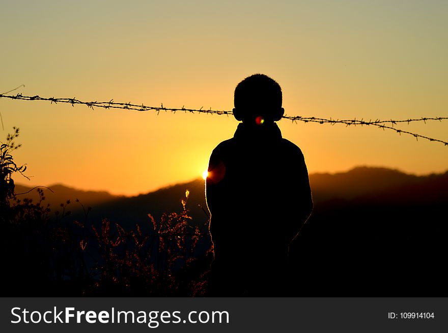 Silhouette of Boy Standing Near Barbed Wire Fence during Golden Hour