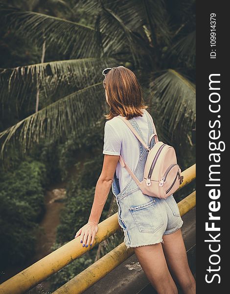 Woman Wearing Dungaree Shorts Stands Near a Yellow Metal Rail Overlook a River Belo With Coconut Trees at Daytime