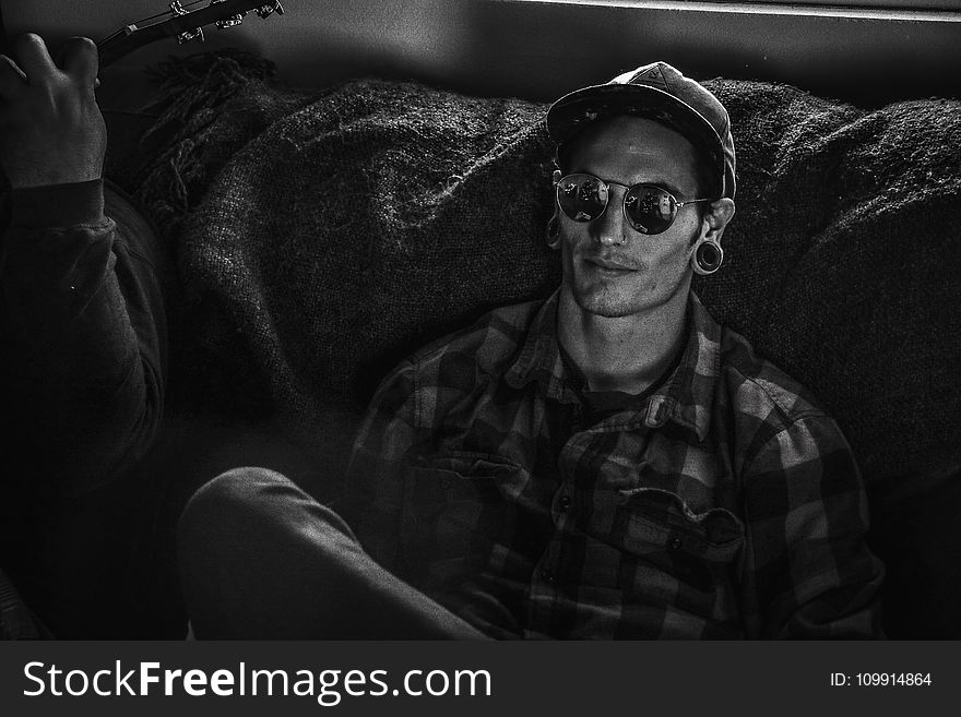 Grayscale Photo Of Man Wearing Snapback Cap And Plaid Dress Shirt Sitting On Couch