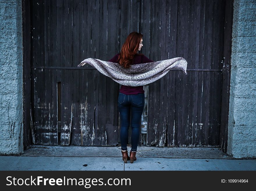 Woman Holding Gray Shawl While Spreading Her Arms Infront Of Brown Wooden Door