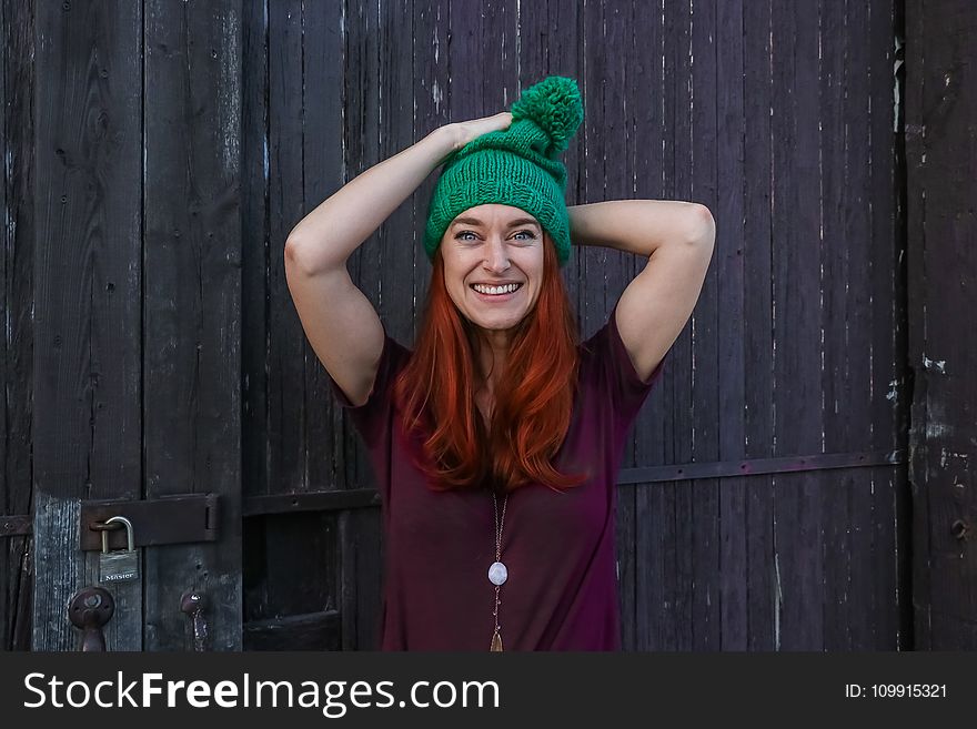 Woman in Green Bobble Cap and Purple T-shirt Posing Near Black Wooden Fence