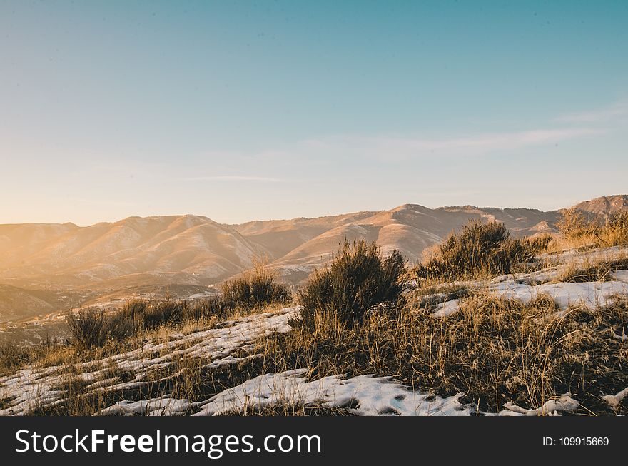 Green And Brown Grass Covered With Snow Overlooking Brown Hills And Mountains Under Clear Blue Sky At Daytime