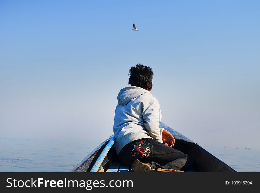 Man in Grey Hoodie and Black Pants Sitting in the Middle of Boat Looking at Bird in the Sky