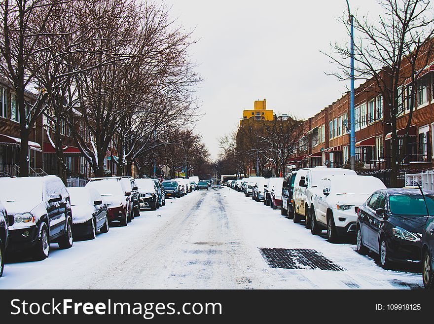 Snow Covered Road and Inline Parked Vehicles Between 2-storey Buildings Under White Sky