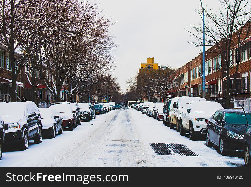 Snow Covered Road And Inline Parked Vehicles Between 2-storey Buildings Under White Sky