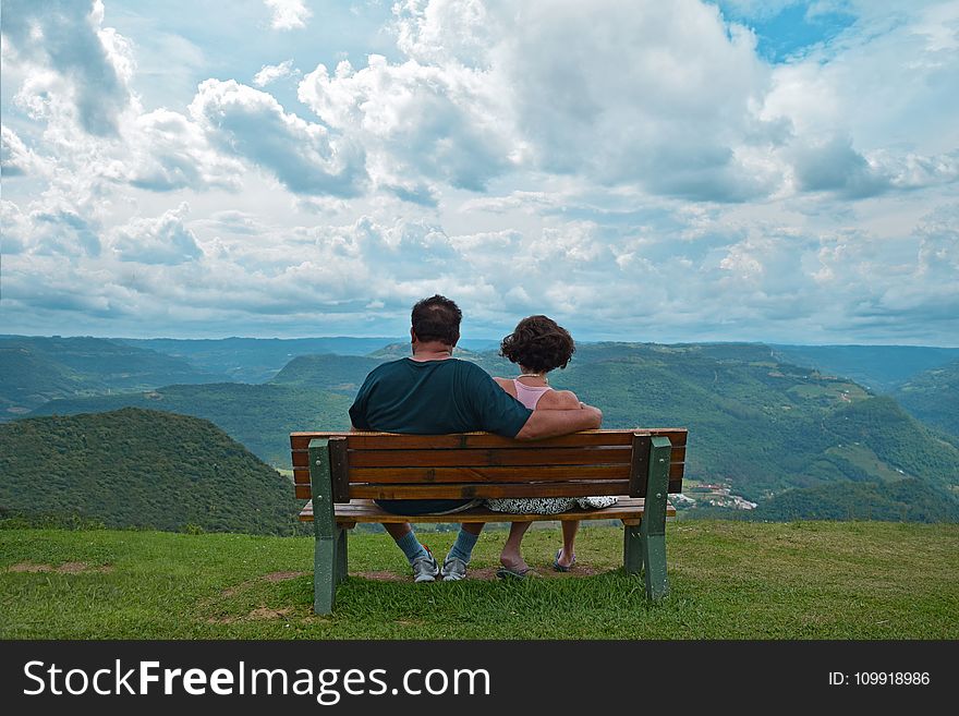 Couple Sitting on Brown Wooden Bench Near Mountains Covered With Grasses Under Blue Cloudy Sky