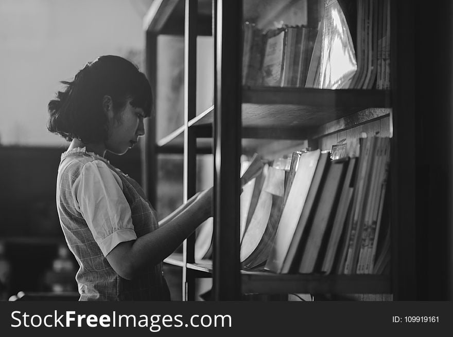 Grayscale Photo of a Woman Holding a Book Inside the Library