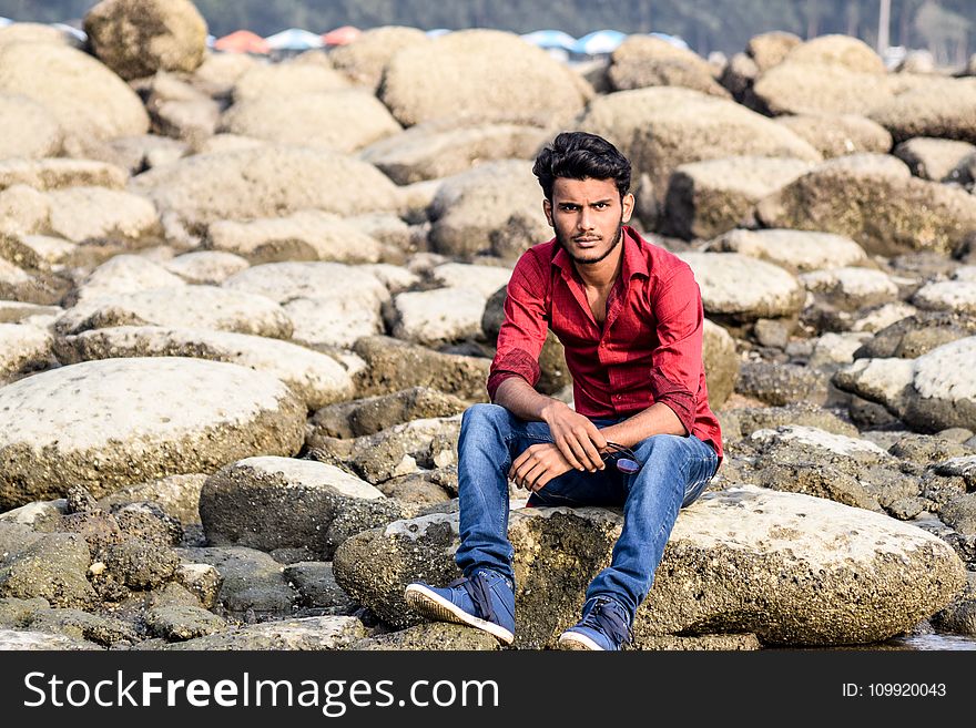Man Wearing Red Dress Shirt and Blue Jeans White Sitting on Gray Stone Formation