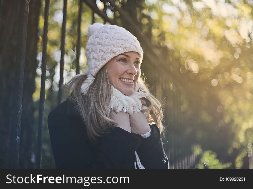 Photography of Woman in Black Jacket and White Knit Cap Smiling Next to Black Metal Fence