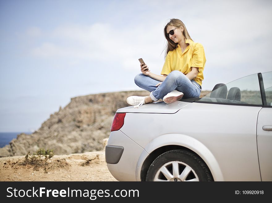 Woman in Yellow Blouse and Blue Jeans Taking Selfie While Sitting on Car