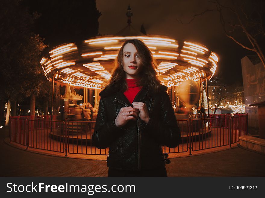 Woman Wearing Black Leather Jacket and Red Turtle-neck Shirt Standing in Front of Carousel Ride