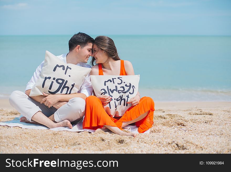 Man and Woman Wearing Cloths Sitting on Brown Sand Near Seashore