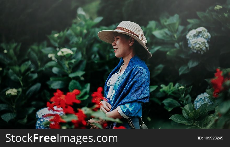 Selective Focus Photo of Woman in Blue Shawl and Brown Sun Hat in the Middle of Garden