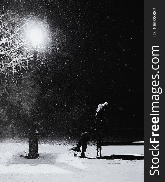 Grayscale Photography of Person With Knit Pompom Cap Sits in Front Turned on Light Post at Night