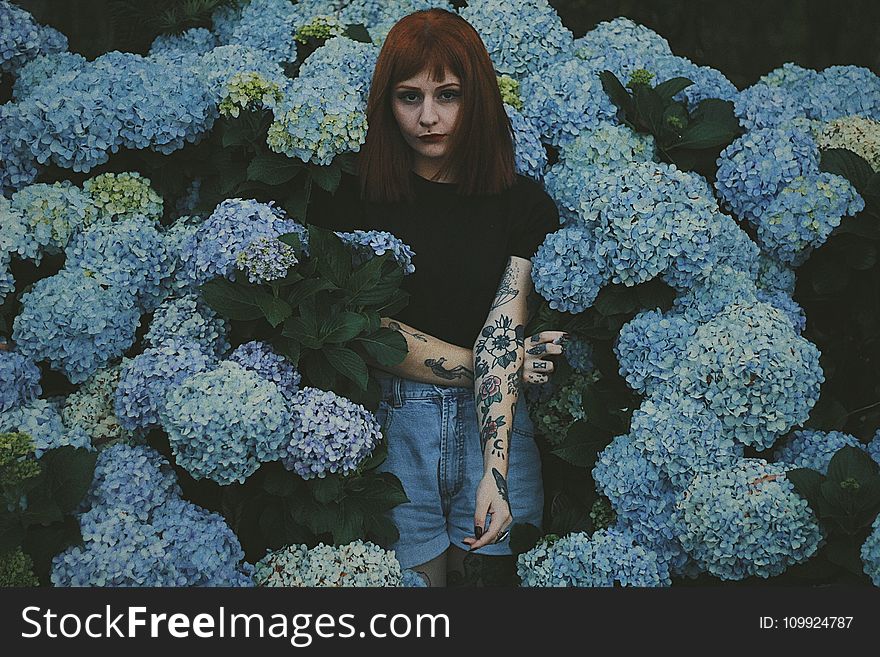 Woman With Black And Red Flower Tattoo Standing Behind Blue Flowers