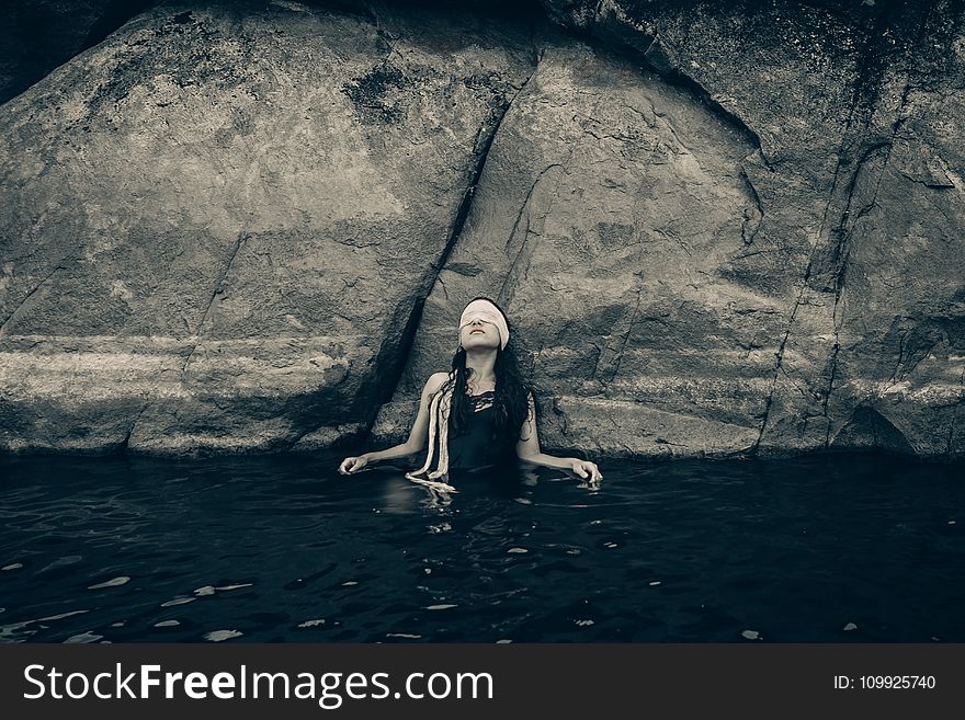 Woman in Blindfold Wearing Black Top on Body of Water While Leaning on a Rock