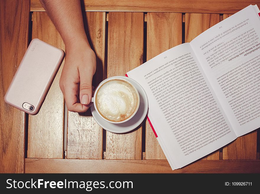 Person&x27;s Hand Holding White Coffee Mug With Plate On Brown Wooden Board With White And Black Book