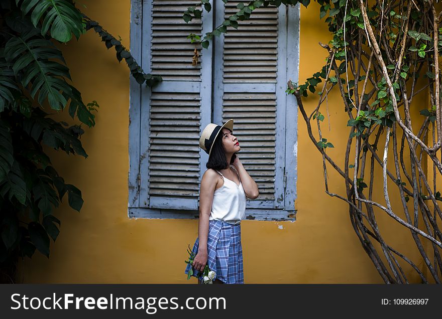 Photo of Woman Wearing White Sleeveless Top and Hat Holding Flowers Near Window