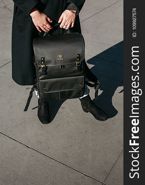Person Wearing Black Coat Holing Black Leather Knapsack Backpack Standing on Concrete Ground