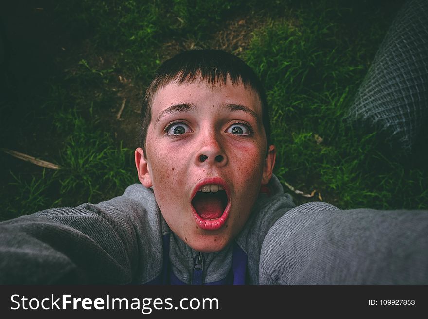 Opened Mouth Black Haired Boy In Gray Full-zip Jacket Standing On Grass Field Taking Selfie