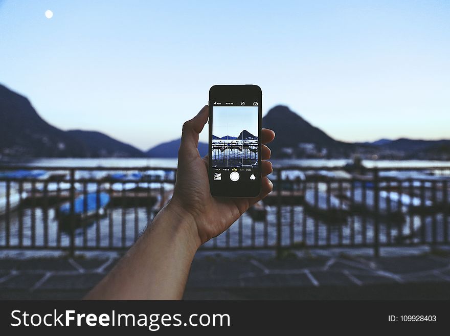 Person Holding Iphone Taking Picture Of Mountain Near Body Of Water