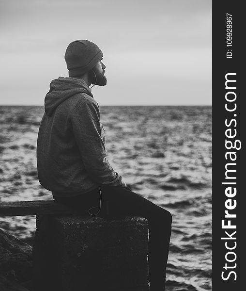 Grayscale Photo of Man in Hoodie and Kit Cap Sitting Near Bodies of Water