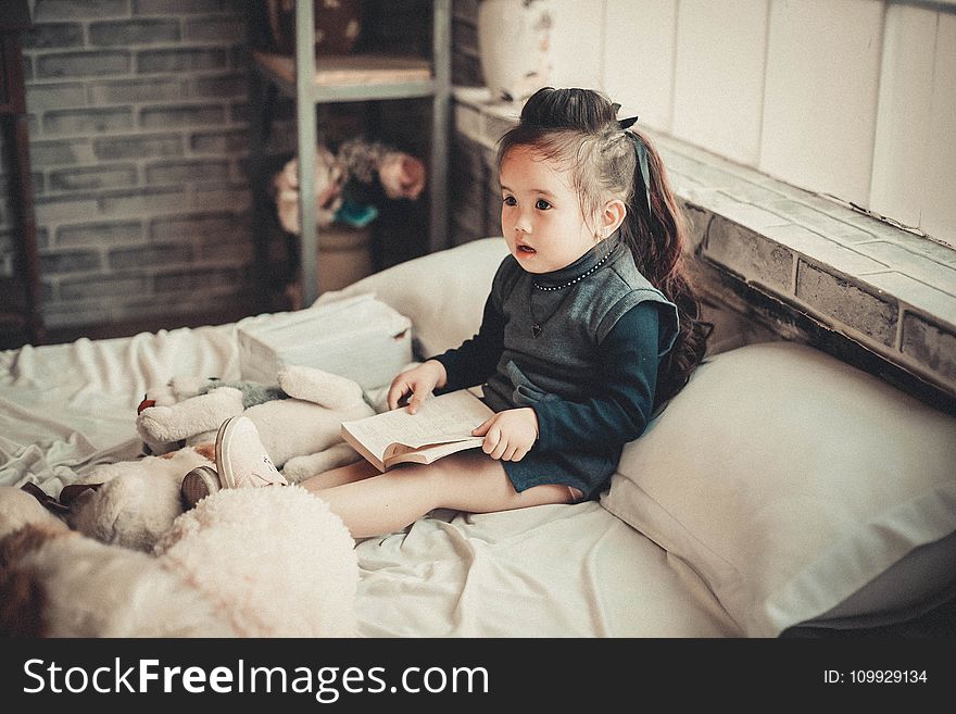 Toddler Girl Wearing Long-sleeved Top Reading Book While Sitting on Bed