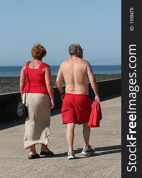 Rear view of a middle aged couple both wearing red, walking together along a beach promenade with the sea and a blue sky to the rear. Rear view of a middle aged couple both wearing red, walking together along a beach promenade with the sea and a blue sky to the rear.